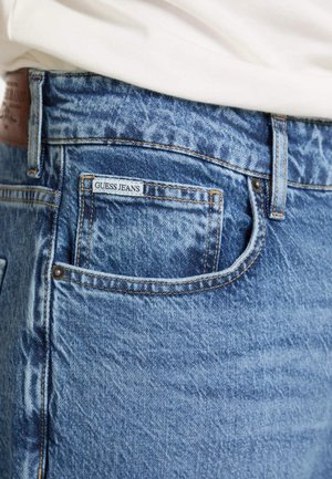 Wyblakłe niebieskie dżinsy zbliżeniowym ujęciem pokazującym metkę Guess Jeans na tylnej kieszeni oraz kontrastowe przeszycia wzdłuż szwów.