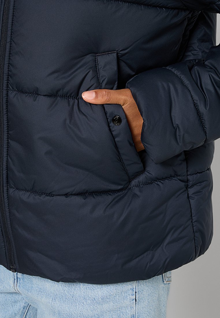 Marineblaues Puffer-Jacke mit gestepptem Muster, Frontreißverschluss, seitlichen Taschen und einem Druckknopfakzent in der Nähe der Öffnung, getragen über hellen Jeans.