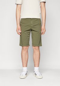 Pantaloni corti di cotone verde oliva con taglio dritto, due tasche frontali e orlo cucito. Indossati con sneakers bianche e calzini.