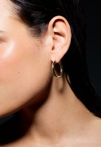 Orecchino a cerchio d'oro, forma circolare, finitura lucida, appeso al lobo. Primo piano di un orecchio con una texture della pelle liscia e capelli scuri.