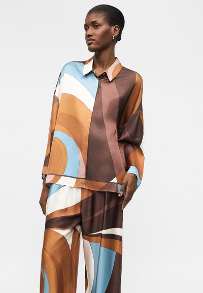 Γυναίκα που φοράει φαρδιά, πολύχρωμη πουκάμισο και παντελόνια με αφηρημένα κυρτά μοτίβα σε καφέ, μπλε, λευκό και μπεζ, όρθια μπροστά σε απλό φόντο.