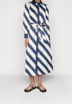 Mujer con un vestido de botones de manga larga, a rayas diagonales en azul marino y blanco, con cinturón en la cintura, combinado con sandalias blancas de tiras sobre un fondo liso.
