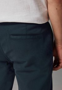 Pantalones oscuros de color azul con una textura suave, que presentan un solo bolsillo trasero y detalles de costura sutiles. Llevados con una camisa de color claro.