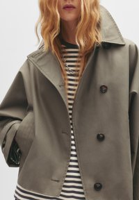 Khaki oversize jakna s širokim ovratnikom, s dva crna gumba i dva skrivena kopča. Ispod se nalazi majica s crno-bijelim prugama.