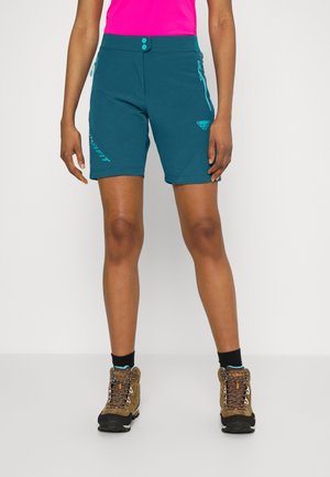 Vrouw draagt teal buitenkorte broek met ritspockets, zwarte sokken, bruine wandelschoenen en een felroze top, staand tegen een witte achtergrond.