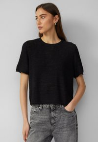 Czarna, dziana bluzka z krótkim rękawem i okrągłym dekoltem, o luźnym kroju i fakturowanym materiale. W zestawie z szarymi, wysokogatunkowymi dżinsami.