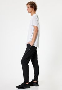 Hvid kortærmet T-shirt med sidebesætninger, parret med sorte tapered joggers med lynlåslommer og elastiske manschetter. Sorte sportsko.