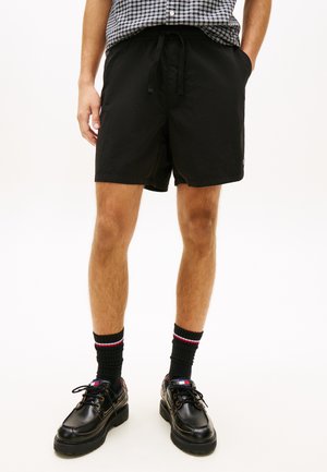 Uomo che indossa pantaloncini neri con coulisse, calzini neri con strisce rosse e bianche e scarpe di pelle nere con lacci, in piedi con le mani in tasca.