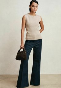 Beige ärmelloses Strickoberteil mit geripptem Saum, kombiniert mit dunkelblauem, ausgestelltem Jeans. Das Model hält eine kleine dunkelbraune Wildlederhandtasche.