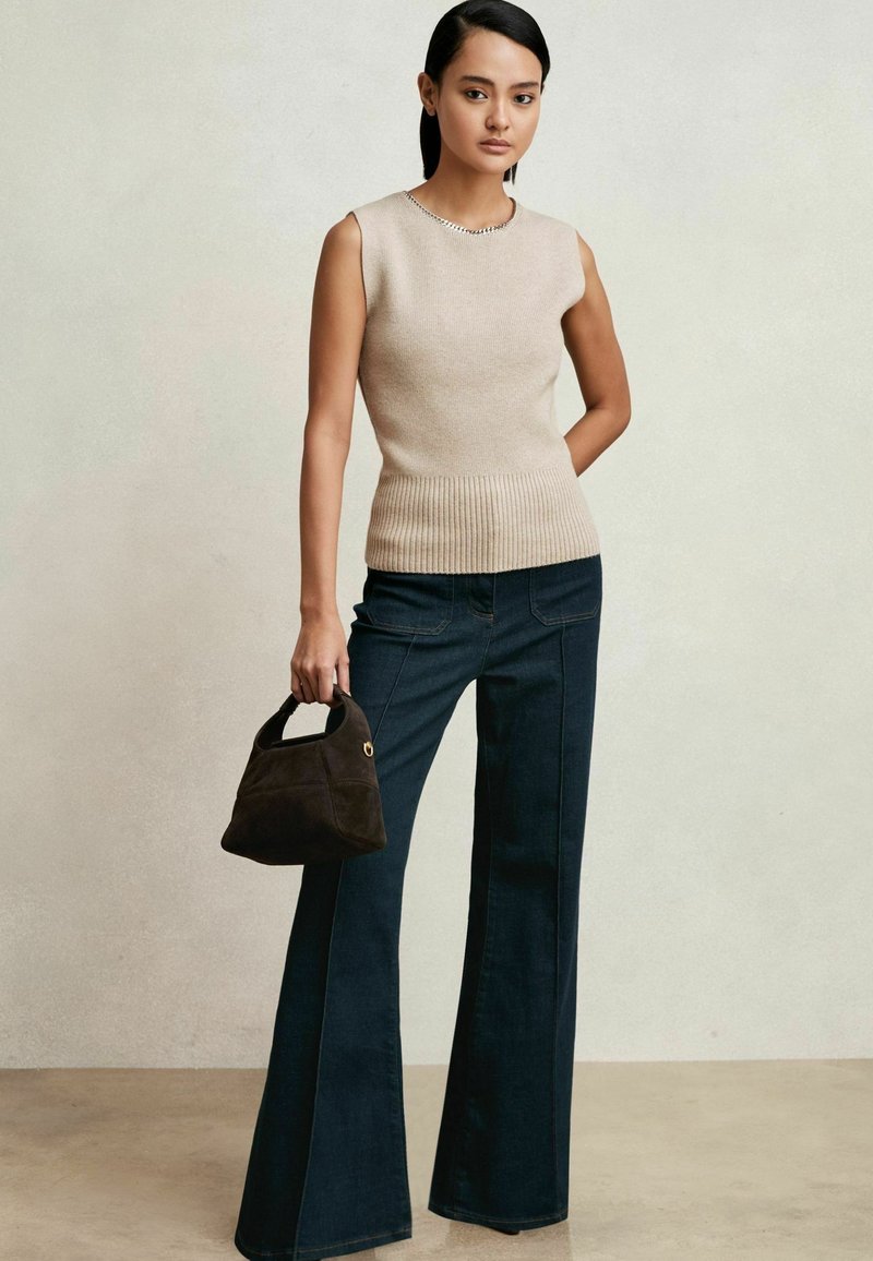 Beige ärmelloses Strickoberteil mit geripptem Saum, kombiniert mit dunkelblauem, ausgestelltem Jeans. Das Model hält eine kleine dunkelbraune Wildlederhandtasche.