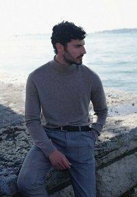 Maglione a collo alto marrone chiaro e pantaloni a quadri grigi. Il tessuto appare morbido con un design aderente, indossato con una cintura nera, seduto vicino all'acqua.