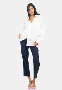 Camicia bianca oversize con bottone, dotata di tasca frontale, abbinata a pantaloni tapered blu navy e tacchi a punta rosa, su uno sfondo neutro.