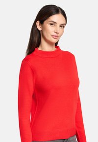 Kobieta z długimi, ciemnymi włosami, ubrana w jasnoczerwoną, długorękawową bluzę, stojąca na tle jednolitego, jasno szarego tła.