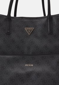 Zwarte tote bag van gestructureerd materiaal. Bevat een gouden driehoekig logo met "GUESS" en een ritscompartiment aan de onderkant.