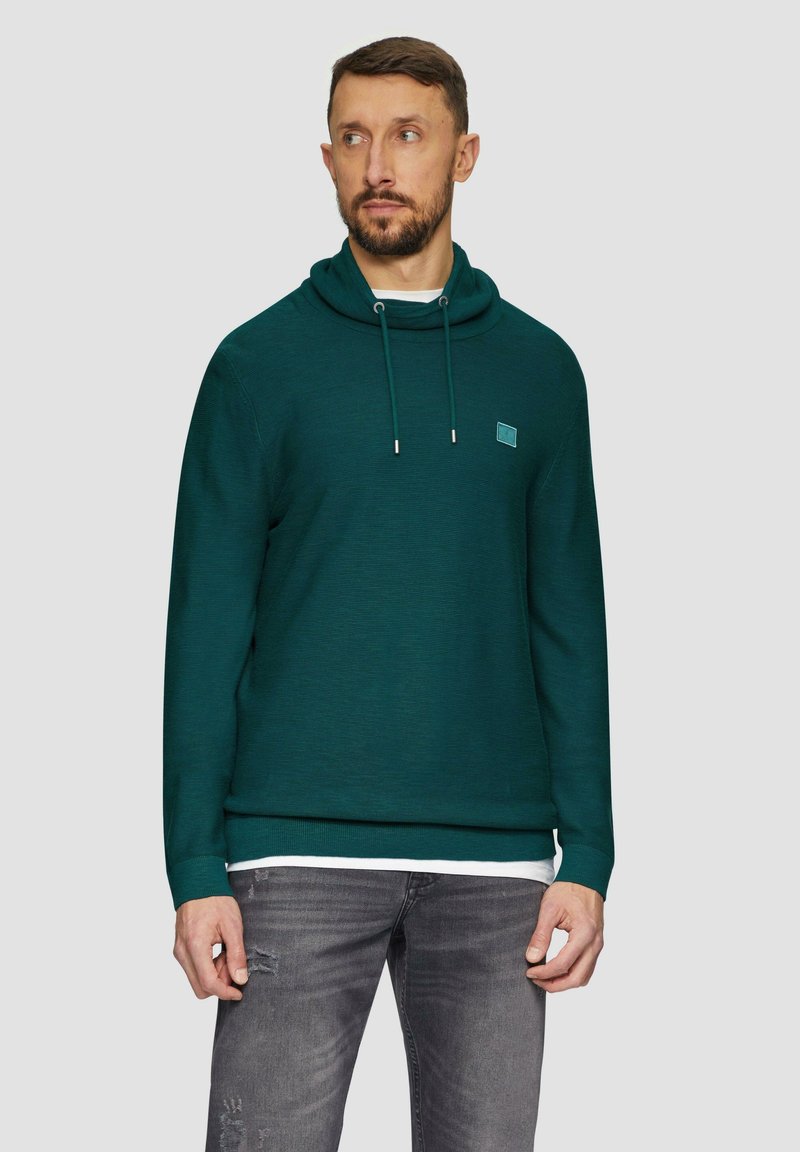 Turkusowy zielony sweatshirt z kapturem, wykonany z teksturowanej tkaniny, z regulowanymi sznurkami i małą łatą z logo na piersi. Połączony z szarymi, przetartymi dżinsami.
