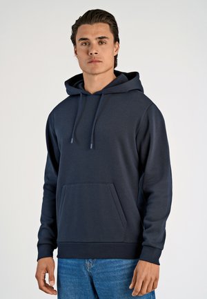 Navyblauwe hoodie van zachte stof, met een kangaroezak, trekkoorden en geribbelde manchetten. Gerdragen met blauwe jeans.