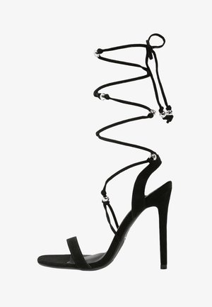 Sandalia de tacón alto de ante negro con finas correas en el tobillo, cuentas decorativas plateadas y un diseño elegante de punta abierta. Cuenta con un tacón de aguja.