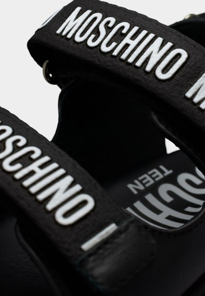 Κοντινό πλάνο μαύρων σανδαλιών με λευκή κεντημένη επωνυμία MOSCHINO σε ρυθμιζόμενα λουράκια και τυπωμένο κείμενο στη σόλα.