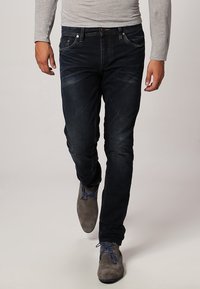 Vaqueros de mezclilla azul oscuro con un corte ajustado, que presentan un sutil desgastado, diseño de cinco bolsillos y herrajes metálicos. Llevados con una camiseta de manga larga gris claro y zapatos grises.