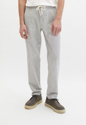 Mann trägt hellgraue Hose mit Kordelzug und dunkelbraune Freizeitschuhe vor weißem Hintergrund.