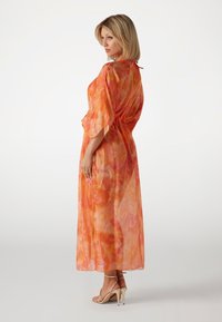 Kaftan lungo e trasparente di un vibrante arancione con motivi floreali. Chiusura a cravatta sul collo, maniche ampie e una silhouette fluida.