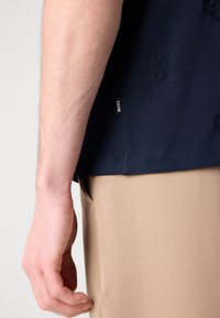 Tamsiai mėlyna trumpų rankovių marškiniai su tekstūruotu raštu, šone esanti maža etiketė 'BOSS' ir matoma smėlio spalvos kelnės medžiaga.