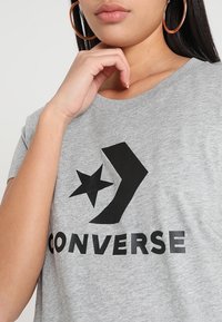 Szara bawełniana koszulka z dużym czarnym logo Converse oraz graficzną gwiazdką na przodzie. Projekt ma klasyczny kształt z okrągłym dekoltem.