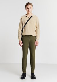 Beige langärmliges Polohemd mit Kragen, kombiniert mit olivgrünen Slim-Fit-Hosen und schwarzen Stiefeletten. Einstellbarer schwarzer Schultergurt inklusive.