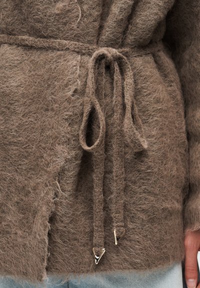 Καφετί πλεκτή ζακέτα με ζώνη δεμένη στη μέση και κορδόνια με μεταλλικές άκρες, σε συνδυασμό με ανοιχτόχρωμο μπλε τζιν, που δείχνει το κάτω μέρος του κορμού και το χέρι.