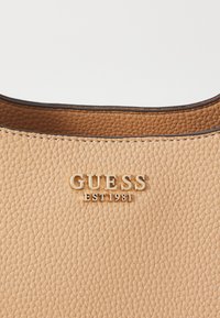 Section de sac à main en cuir texturé beige avec logo doré "GUESS EST 1981" et bordure marron foncé le long du bord supérieur.