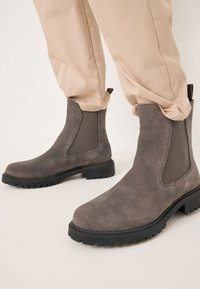 Bottines en daim gris avec panneaux latéraux élastiques et semelle extérieure en caoutchouc noir robuste. Conception simple avec un bout arrondi et des accents minimaux.