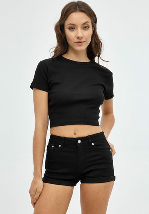 Joven mujer con cabello castaño que lleva una camiseta negra corta y shorts negros de cintura alta sobre un fondo liso.
