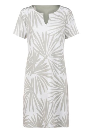 Vrouw met kort blond haar die een witte knielange jurk draagt met grijze palmbladprint en witte sandalen met hak.