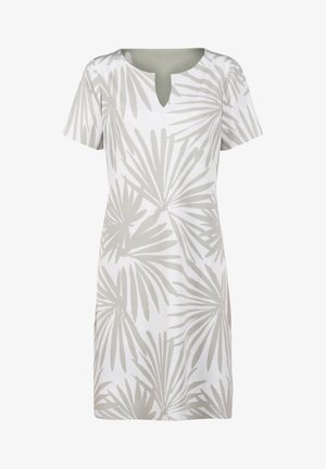 Femme aux cheveux blonds courts portant une robe blanche mi-longue avec un motif feuilles de palmier gris et des sandales blanches à talons.
