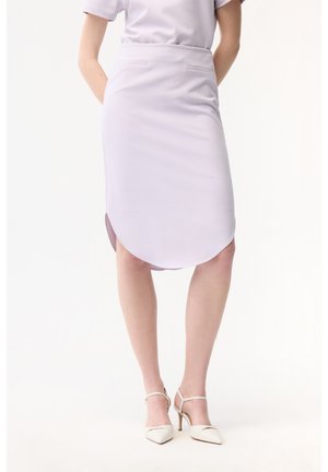 Kvinde iført en lyse-lilla pencilnederdel med afrundet kant og hvide højhælede sko med ankrem, stående med hænderne bag ryggen.