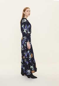 Blumenmuster, langärmliges Kleid aus dunkelblauem Stoff mit grünen und lila Akzenten. Es hat einen gestuften Rock und seitliche Taschen, kombiniert mit schwarzen Knöchelstiefeln.