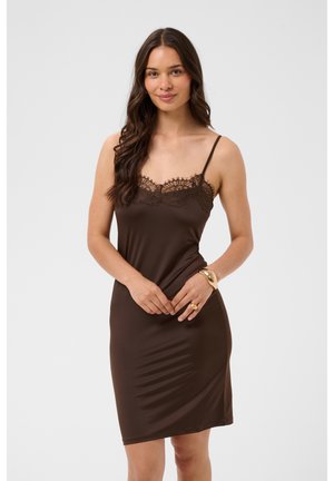 Femme aux longs cheveux foncés porte une robe nuisette marron sans manches avec une bordure en dentelle, debout et souriante devant un fond uni clair.