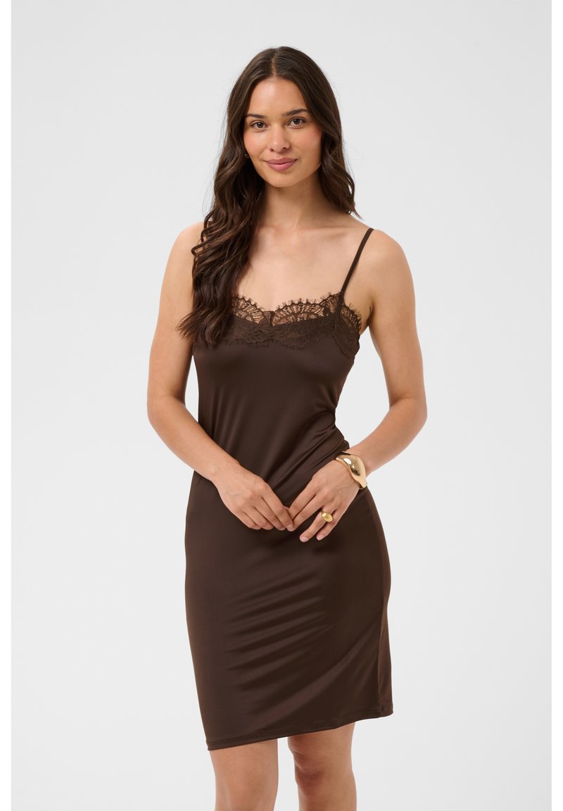 Femme aux longs cheveux foncés porte une robe nuisette marron sans manches avec une bordure en dentelle, debout et souriante devant un fond uni clair.