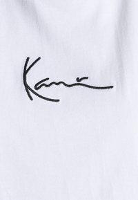 Hvit stoff med en glatt tekstur som har en svart brodert signatur "Kani" i kursiv skrifttype, sentrert og fremtredende.