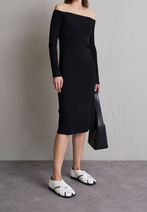 Femme portant une robe midi noire à manches longues et épaules dénudées, tenant un sac cabas noir et portant des sandales blanches à bout fermé.