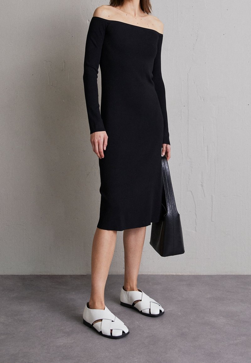 Femme portant une robe midi noire à manches longues et épaules dénudées, tenant un sac cabas noir et portant des sandales blanches à bout fermé.