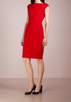 Kobieta w dopasowanej, czerwonej sukience do kolan z marszczeniami i czarnych szpilkach, stojąca na jednolitym beżowym tle.