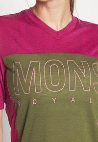 Triko s výstřihem do V v růžové a olivově zelené barvě, s velkým růžovým textem "MONS ROYAL," kontrastními barevnými bloky a měkkou texturou.