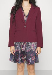 Burgundský blazer s jedním knoflíkem a dvěma předními kapsami, přehozený přes tmavé šaty s květinovými a paisley vzory.