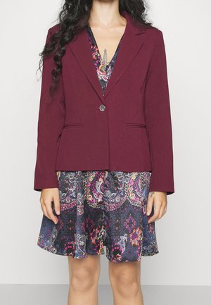Blazer bordeaux à un bouton et deux poches avant, superposé sur une robe sombre à motifs floraux et paisley.