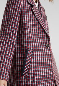 Nærbillede af en skræddersyet frakke med et rød, hvid og blå houndstooth mønster og en enkelt sort knap som lukning nær taljen.