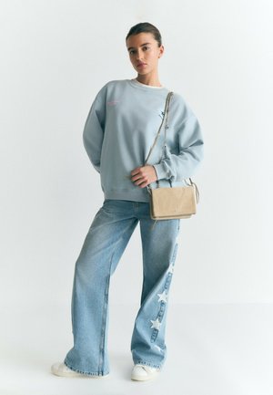 Joven mujer con sudadera azul claro y vaqueros anchos con parches de estrellas, sosteniendo un bolso beige cruzado con correa de cadena.