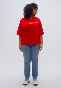 Κόκκινο βαμβακερό μπλουζάκι με λευκό λογότυπο "TOMMY HILFIGER", χαλαρή εφαρμογή, συνδυασμένο με ανοιχτόχρωμο μπλε τζιν και λευκά αθλητικά παπούτσια, μονόχρωμο φόντο.