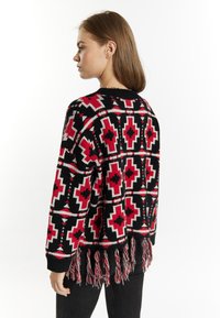 Geweven cardigan in zwart, rood en wit met geometrische patronen en een franjeszoom. Heeft lange mouwen en een ronde hals.