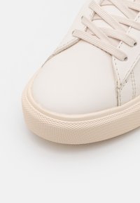 Zapatilla de cuero blanca con puntas redondeadas, cordones planos y suela de goma texturizada con un acento beige. Acabado liso con sutiles detalles de costura.